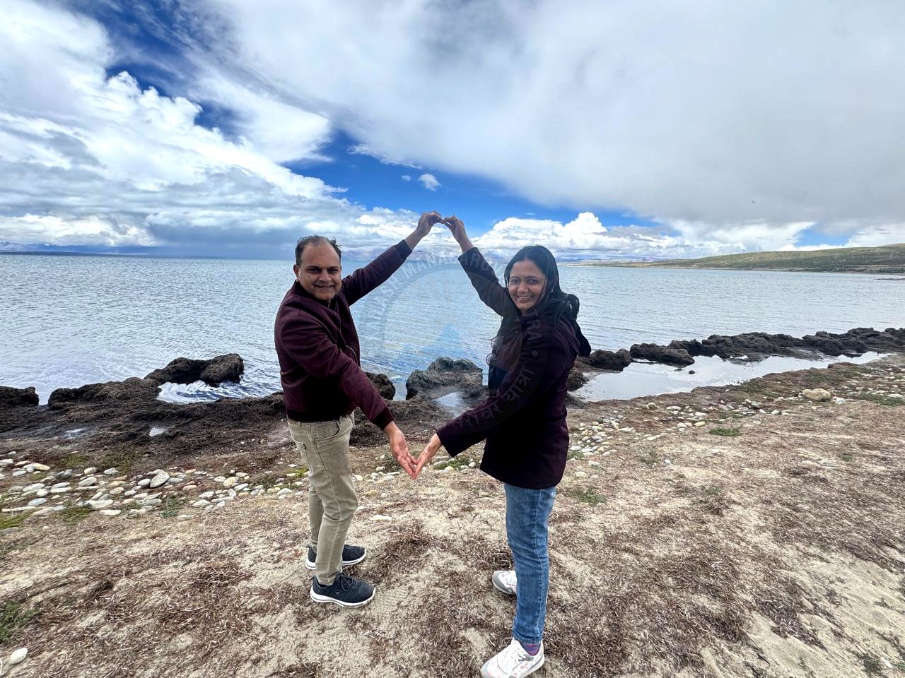 A joyful couple forms a heart with their hands on the serene shores of Lake Tso Moriri in Ladakh, with dramatic clouds and tranquil waters in the background, capturing love and natural beauty.