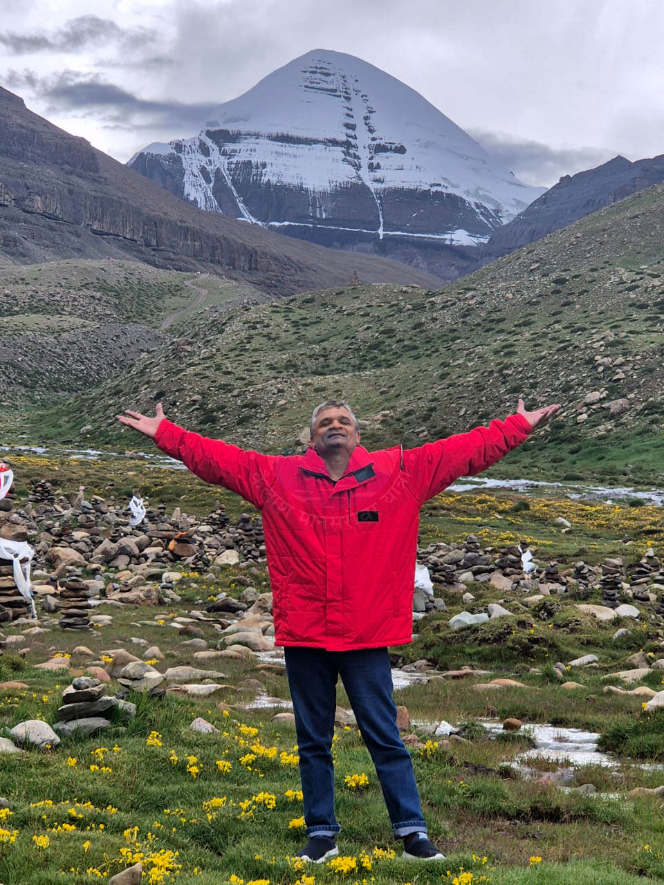 A pilgrim wearing a red jacket stands in green pastures with yellow flowers, beside a stream, with the snow-capped Mount Kailash towering in the cloudy Himalayan sky, radiating joy and a connection to nature.