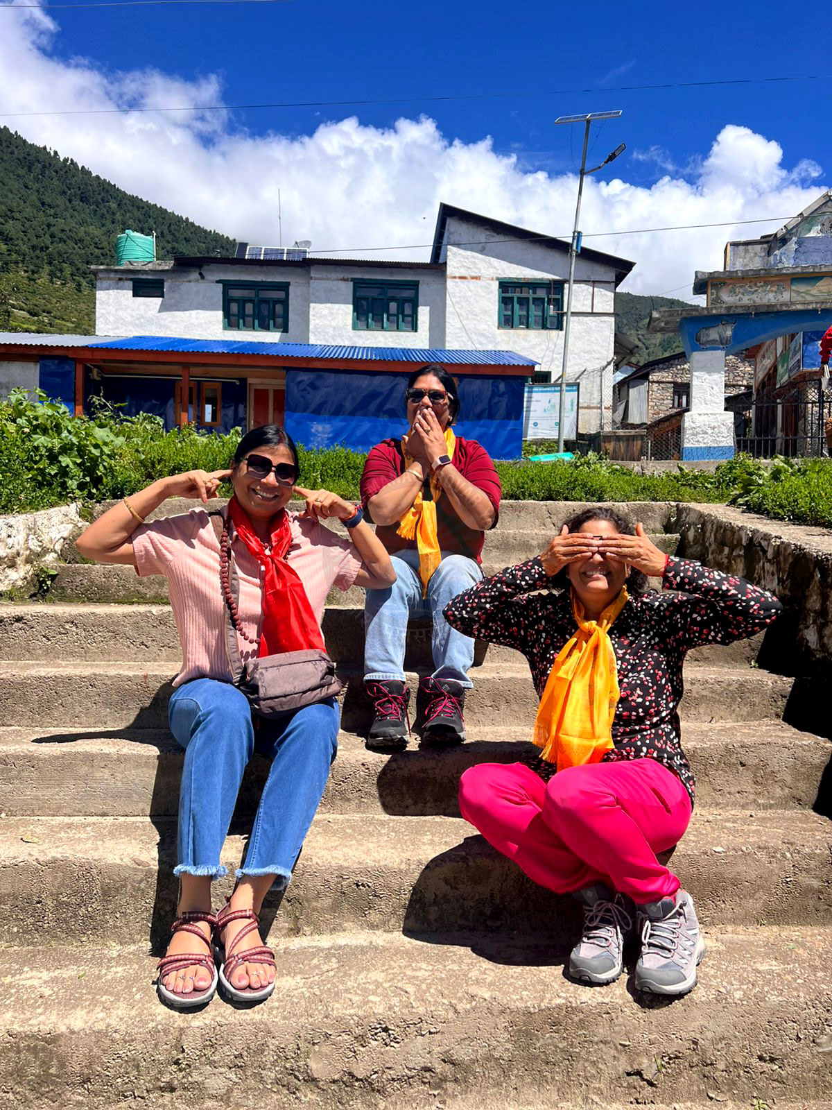Three pilgrims pause on ancient stone steps in the Nepal mountains, wearing bright scarves that symbolize faith and friendship in a peaceful sunlit setting.
