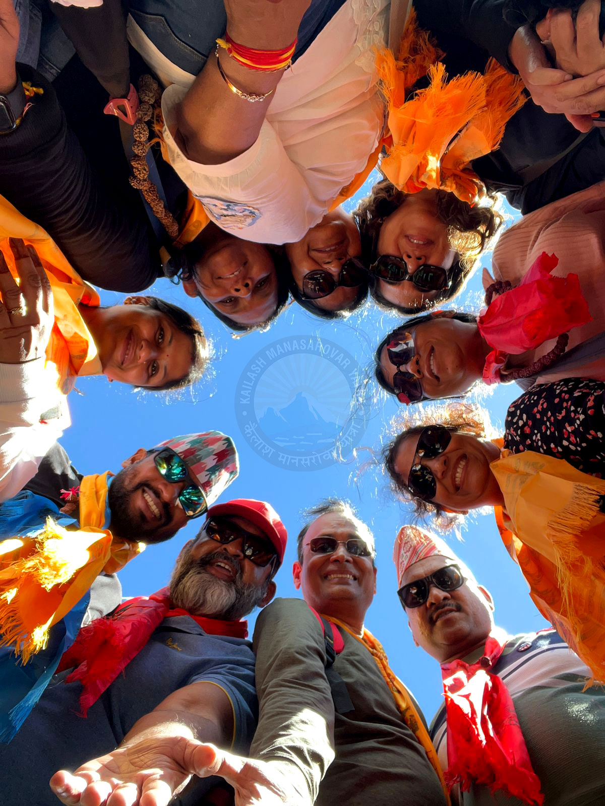 A group of lively pilgrims and friends huddled together, wearing colorful scarves and sunglasses, smiling joyfully against the backdrop of Kailash Manasarovar peak under a clear blue sky, capturing a moment of shared spiritual bliss.