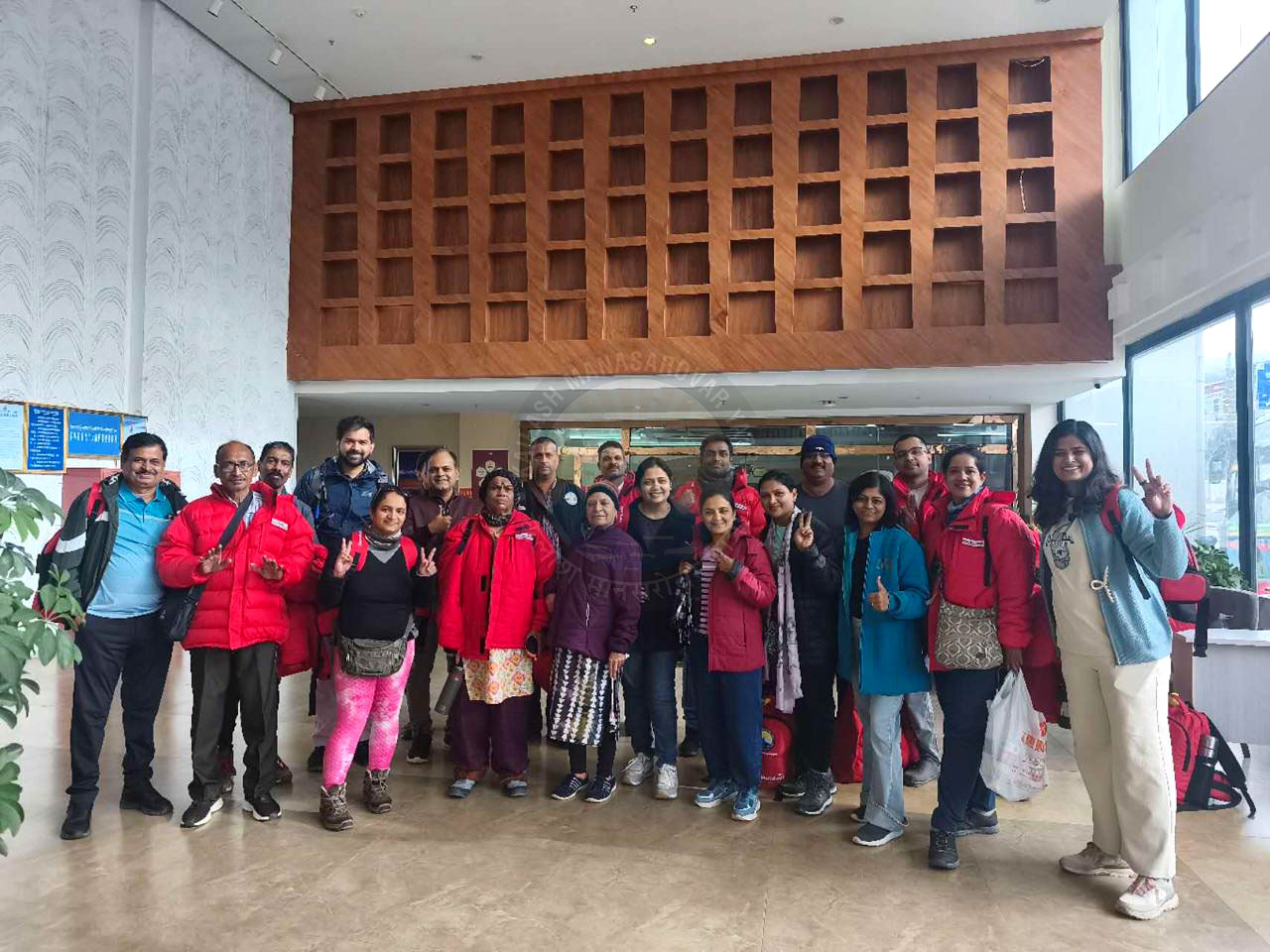 A group of pilgrims in warm jackets posing indoors, smiling and flashing peace signs in a hotel lobby before starting their Kailash Yatra, radiating excitement and spiritual togetherness.
