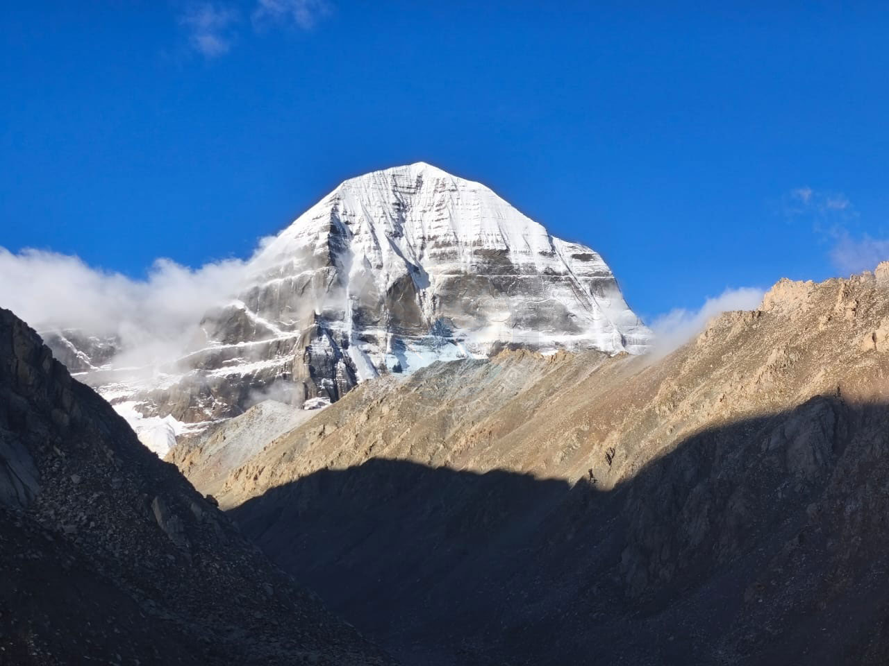 Snow-covered Mount Kailash rising into a clear blue sky, with sunlight casting shadows on surrounding brown mountains and clouds hovering near the lower slopes.