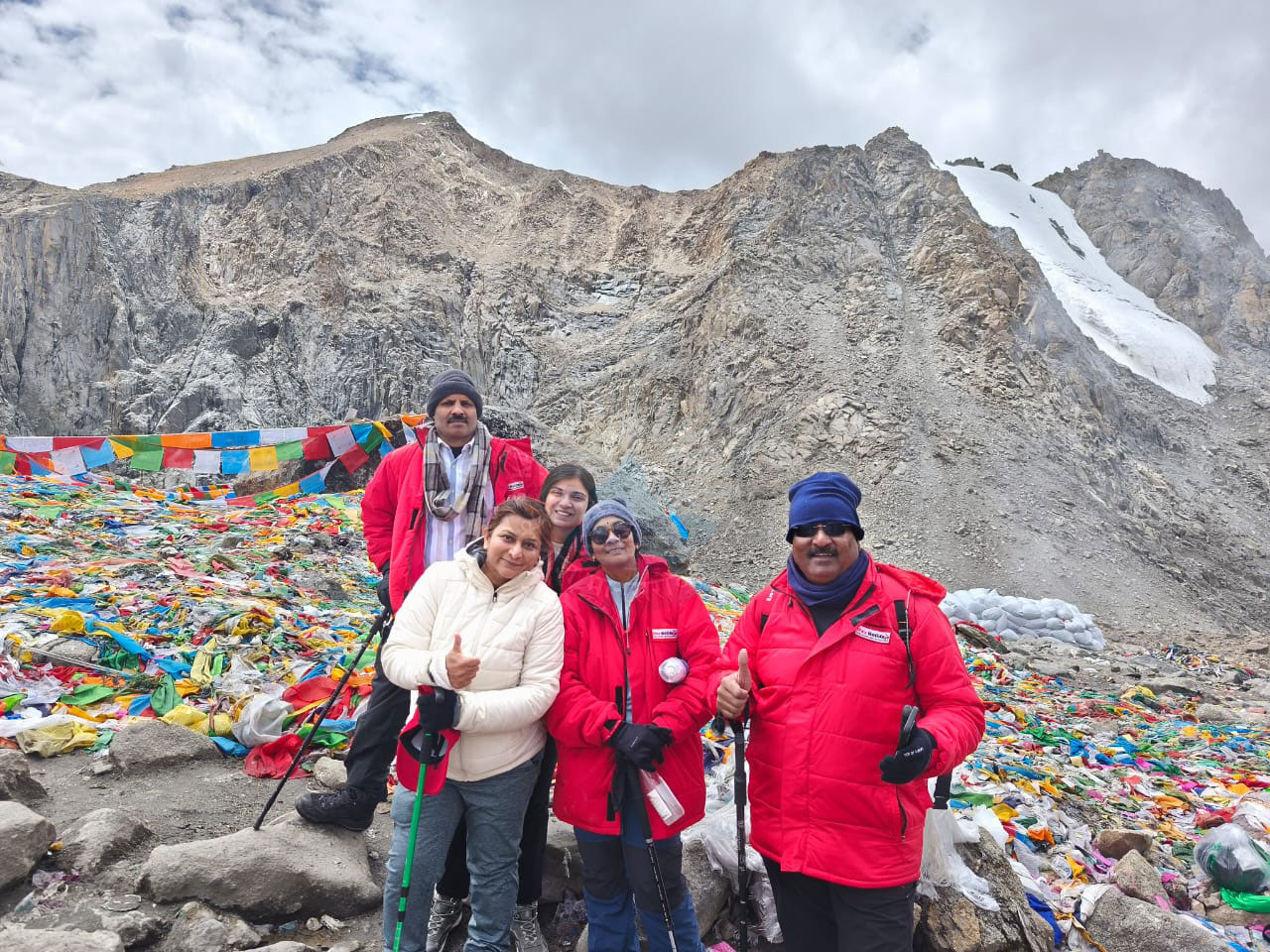 Travelers wearing bright red jackets stand before the craggy, snow-covered peaks of Mount Kailash, surrounded by colorful prayer flags and offerings, capturing the awe and spiritual essence of the Kailash Yatra
