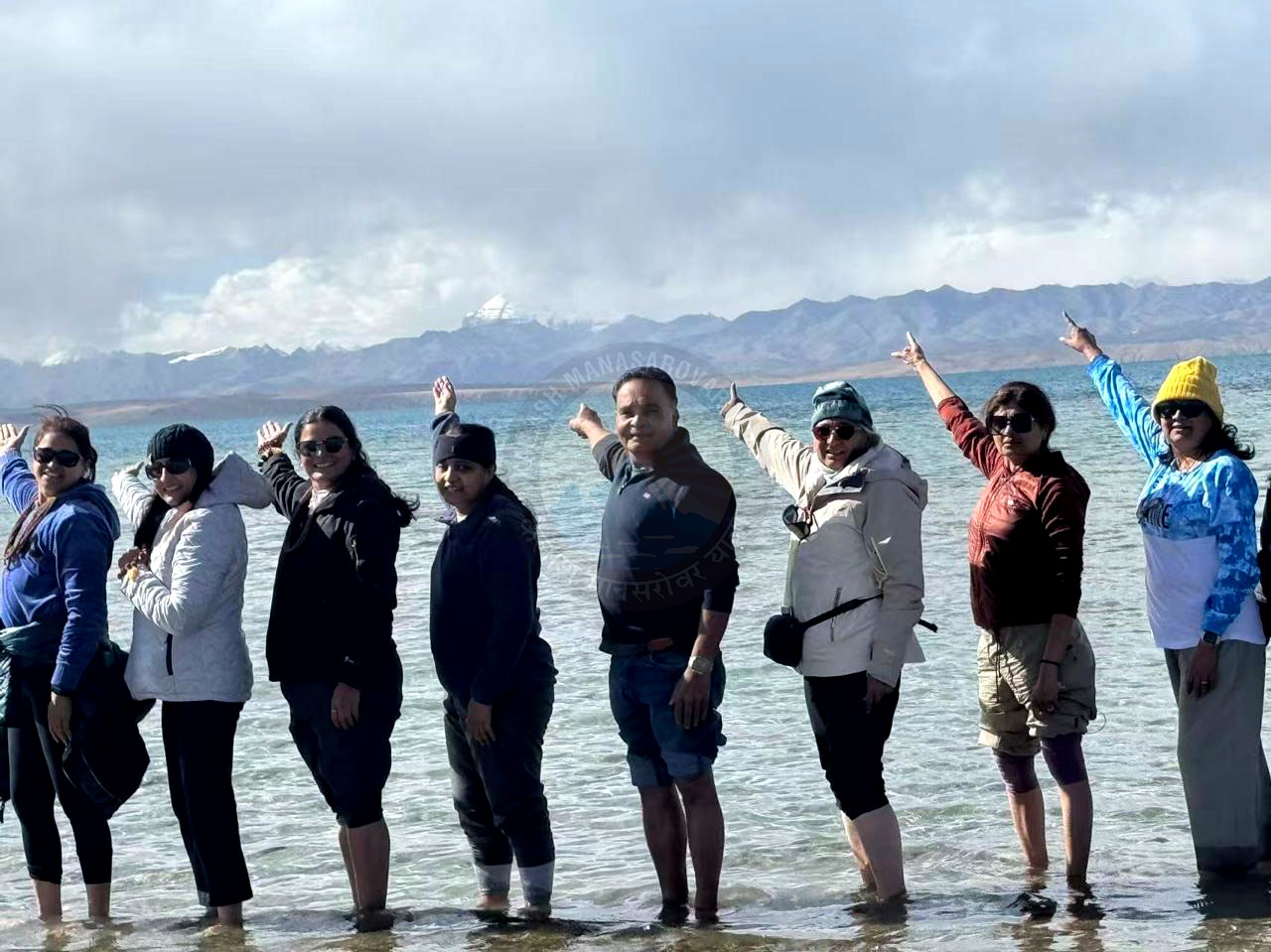A group of pilgrims standing at the edge of Lake Manasarovar, with snow-capped Mount Kailash in the background, enjoying the serene natural and spiritual beauty under a clear sky.
