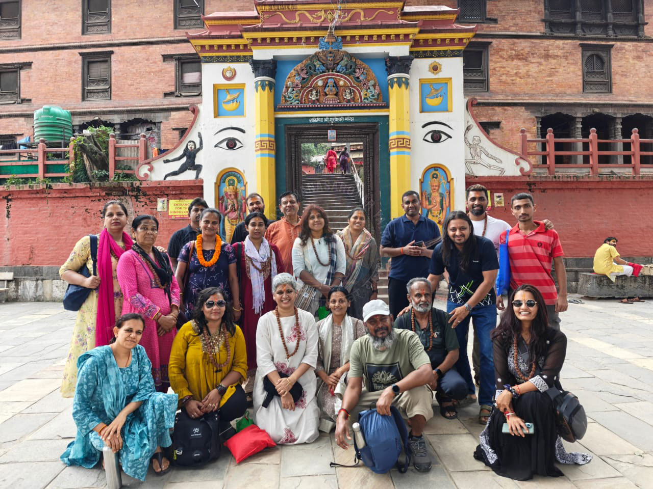 A group of joyful pilgrims in colorful traditional clothing posing in front of a temple entrance in Kathmandu, Nepal, with ornate carvings and historical architecture in the background.
