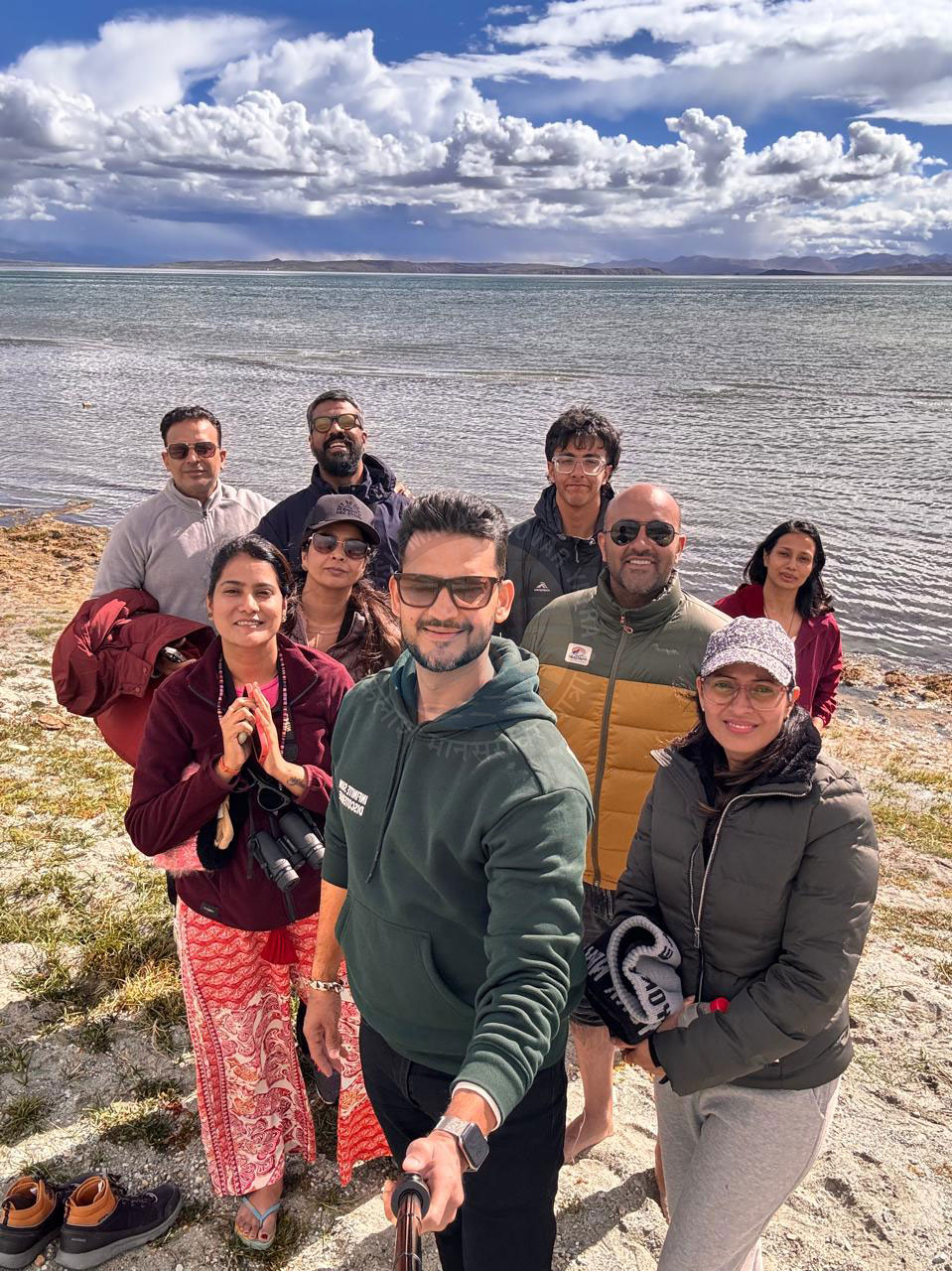 A group of smiling pilgrims posing by the pristine Lake Tso Moriri in Ladakh, with clear blue skies and picturesque clouds in the background.