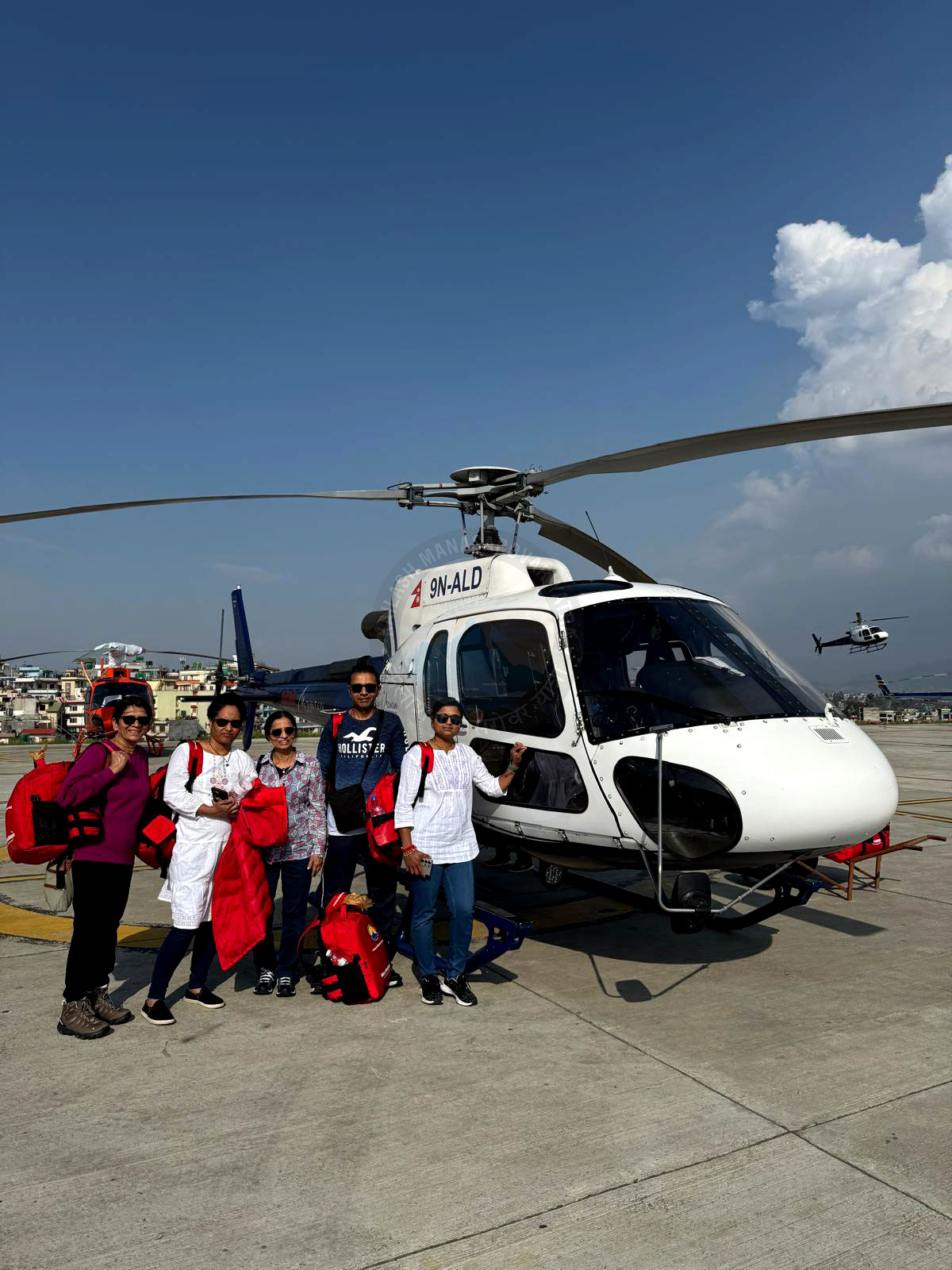 A group of five travelers, including men and women, pose for a photo in front of a white helicopter on a helipad. The group is smiling, and some are wearing life vests and holding bags, with another helicopter visible in the distance.