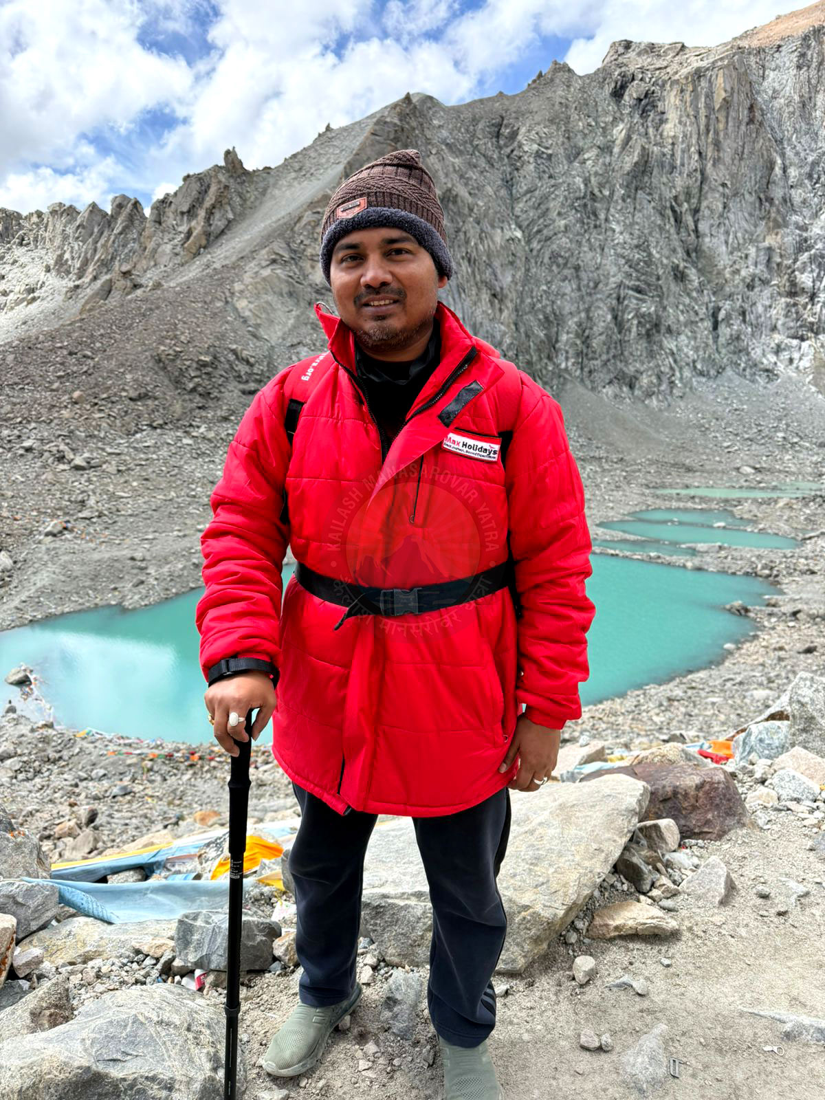 A triumphant hiker wearing a bright red jacket stands on Mount Kailash, holding a walking stick. Behind him, blue lakes shimmer under a grey cloudy sky, capturing a serene sense of accomplishment in a rugged mountainous landscape.