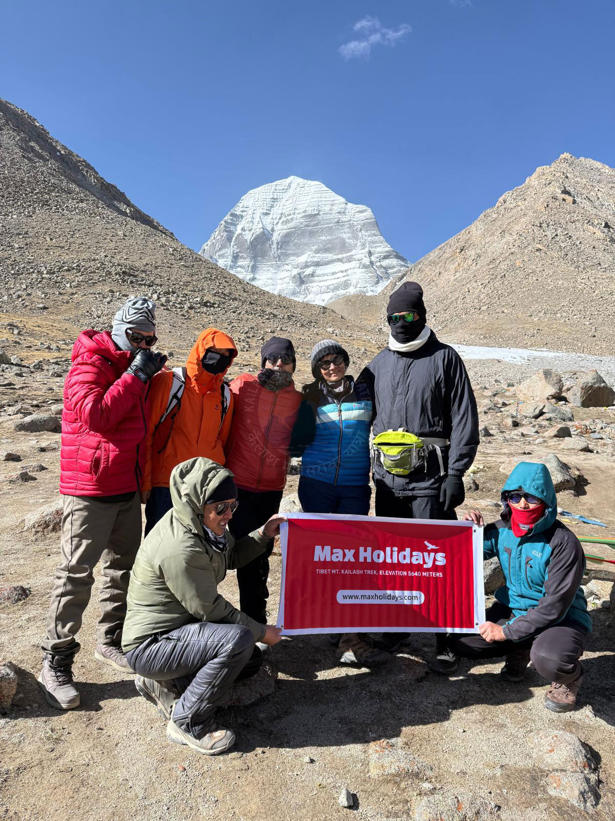 A group of trekkers dressed in heavy clothes hold a 'Max Holidays' sign in front of snow-covered Mount Kailash. The mountain rises dramatically in a pyramid-like shape against a clear blue sky, with rocky, uneven terrain beneath their feet.