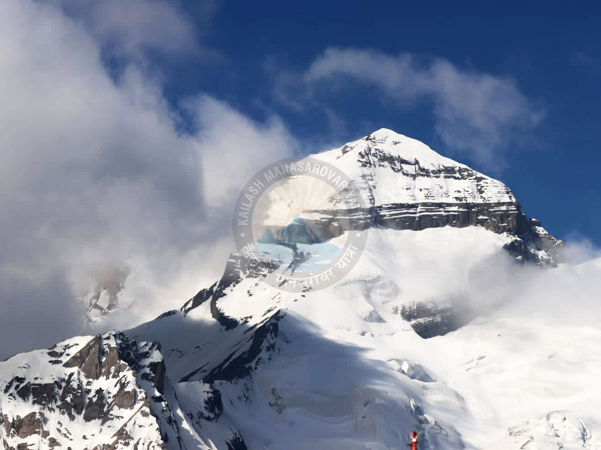 An aerial view of the snow-covered Adi Kailash peak, with a clear blue sky and some white clouds. A watermark for Kailash Mansarovar Yatra is visible on the image.