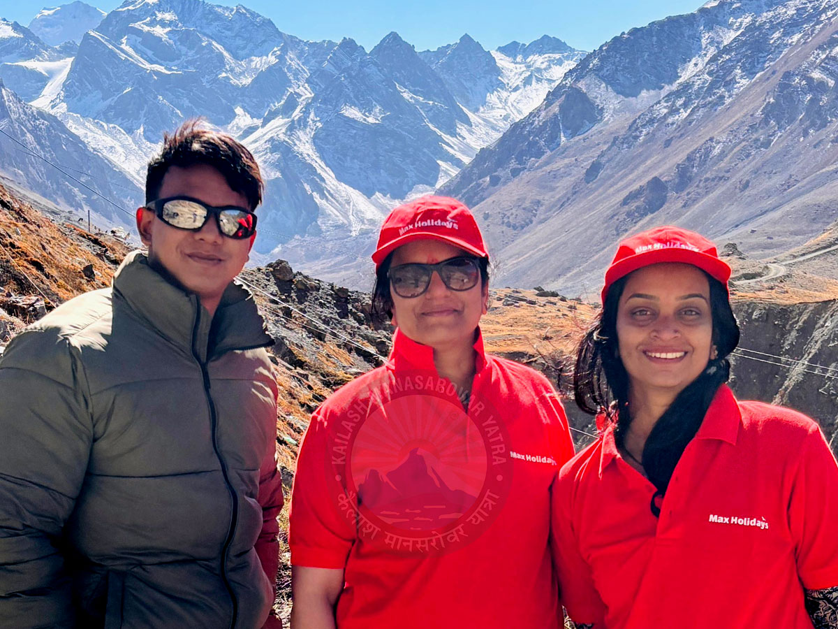 A photo of three smiling trekkers standing in front of the snow-capped Himalayan mountains. The trekkers in the center and on the right are wearing red shirts and hats with Max Holidays printed on them. A watermark for Kailash Mansarovar Yatra is visible in the middle of the image.