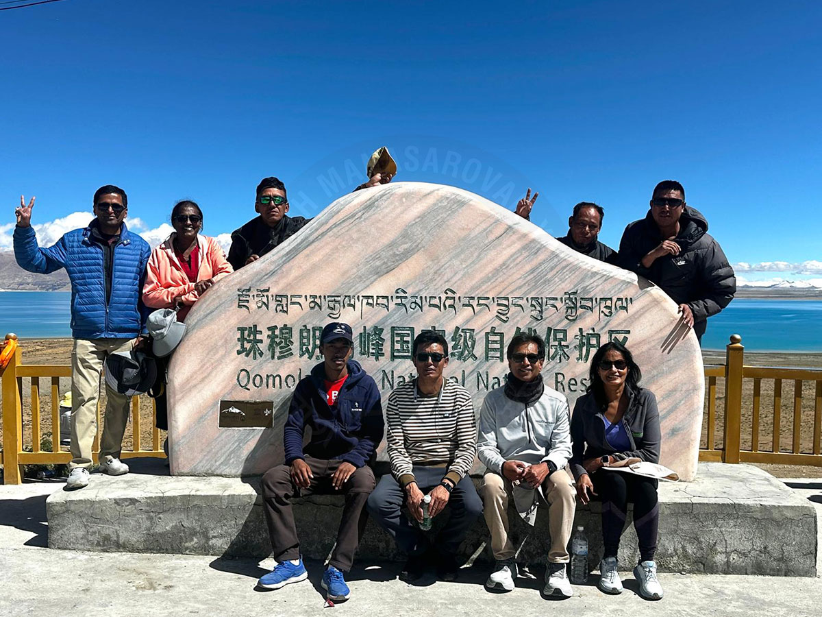 A group of tourists posing in front of a stone sign at Qomolangma National Nature Reserve in Tibet, with a scenic lake and mountain backdrop under a clear blue sky.