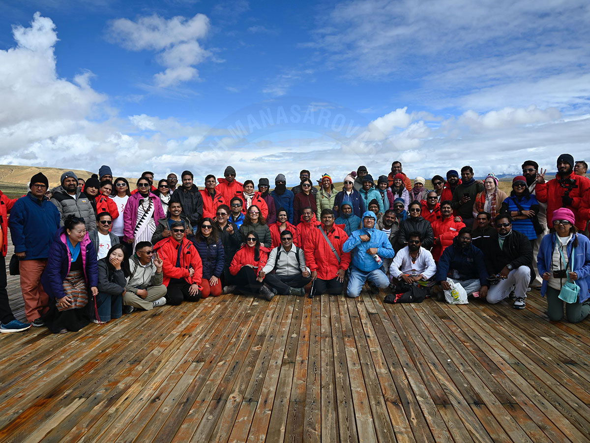 Large group of pilgrims posing on a wooden deck during Mount Kailash and Lake Manasarovar Yatra under a partly cloudy sky