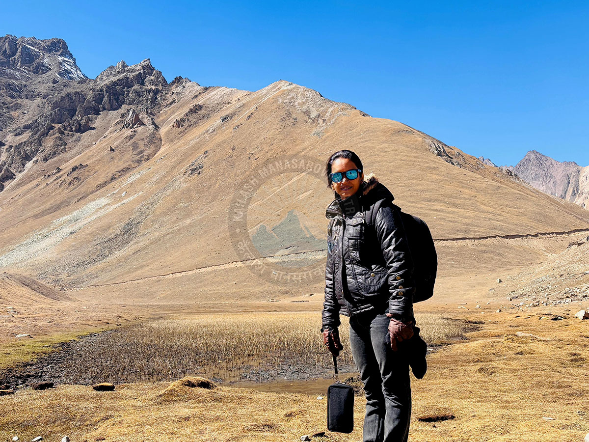 A photo of a woman wearing a black puffer jacket and sunglasses, with a backpack. She is standing in front of a large, dry mountain landscape with a clear blue sky. A watermark for Kailash Mansarovar is visible in the middle of the image.