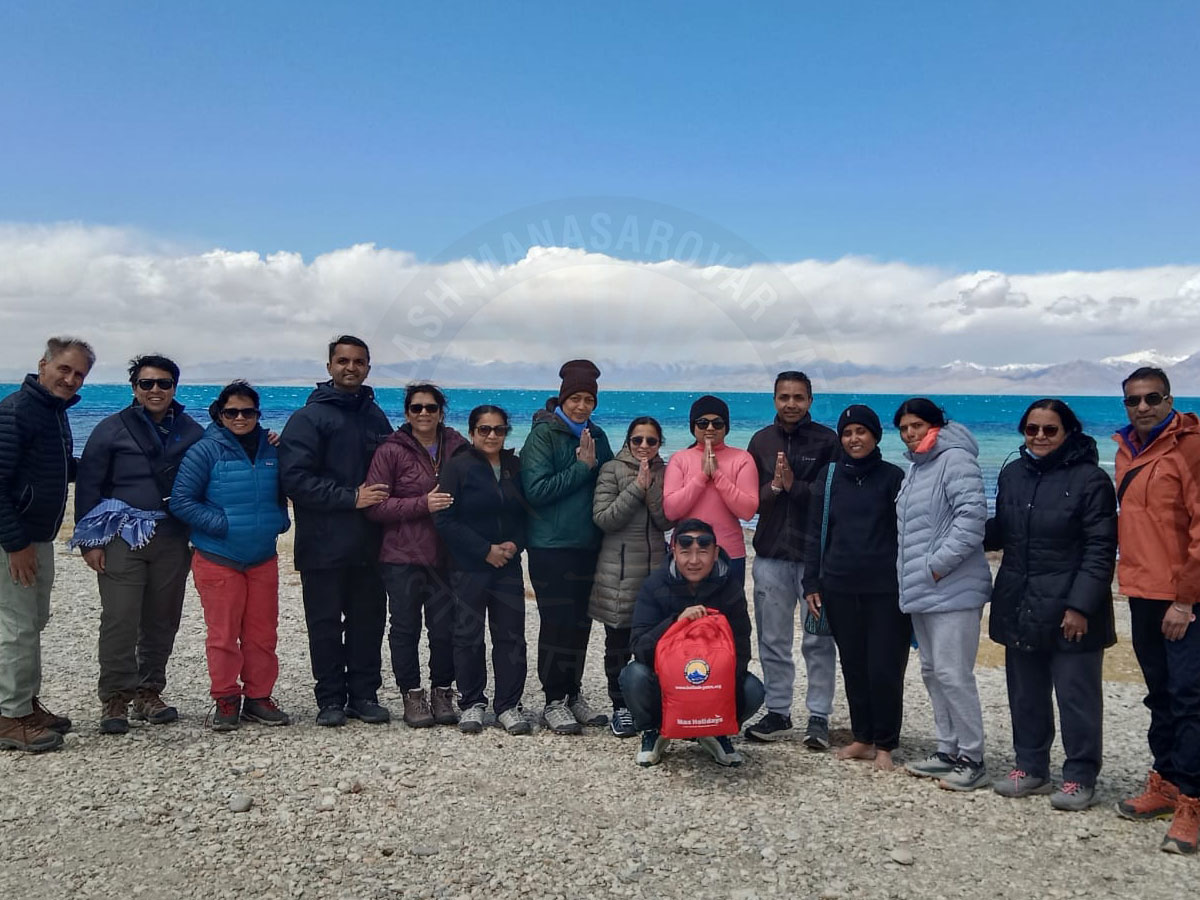 Devotees group photo during Kailash Mansarovar Yatra at Mansarovar Lake with mountains in the background