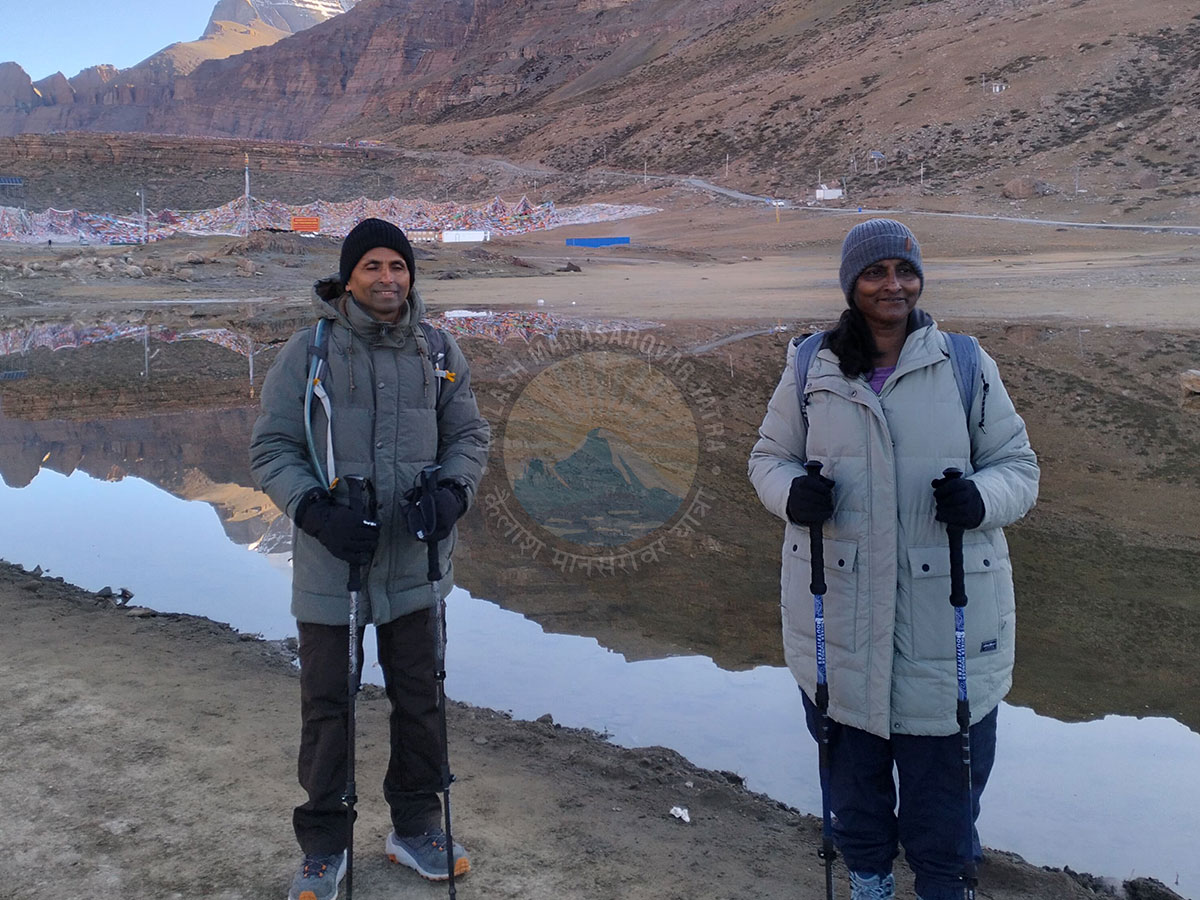 Two pilgrims in warm trekking gear standing beside a clear reflective lake during the Kailash Mansarovar Yatra, with mountains and prayer flags in the background.