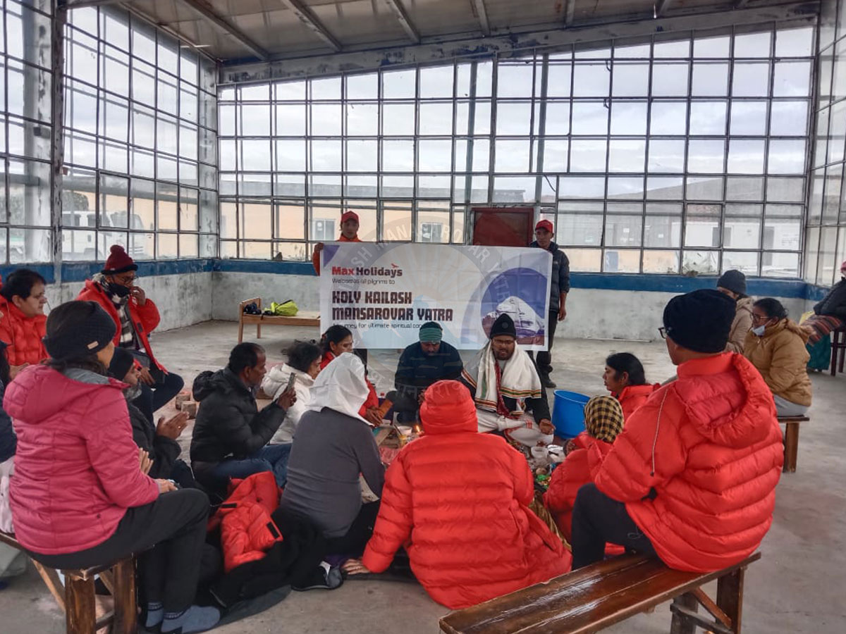 Hindu devotees wearing warm jackets performing a traditional pooja ritual indoors during the Kailash Mansarovar Yatra, with sacred offerings and prayer items placed on a mat.