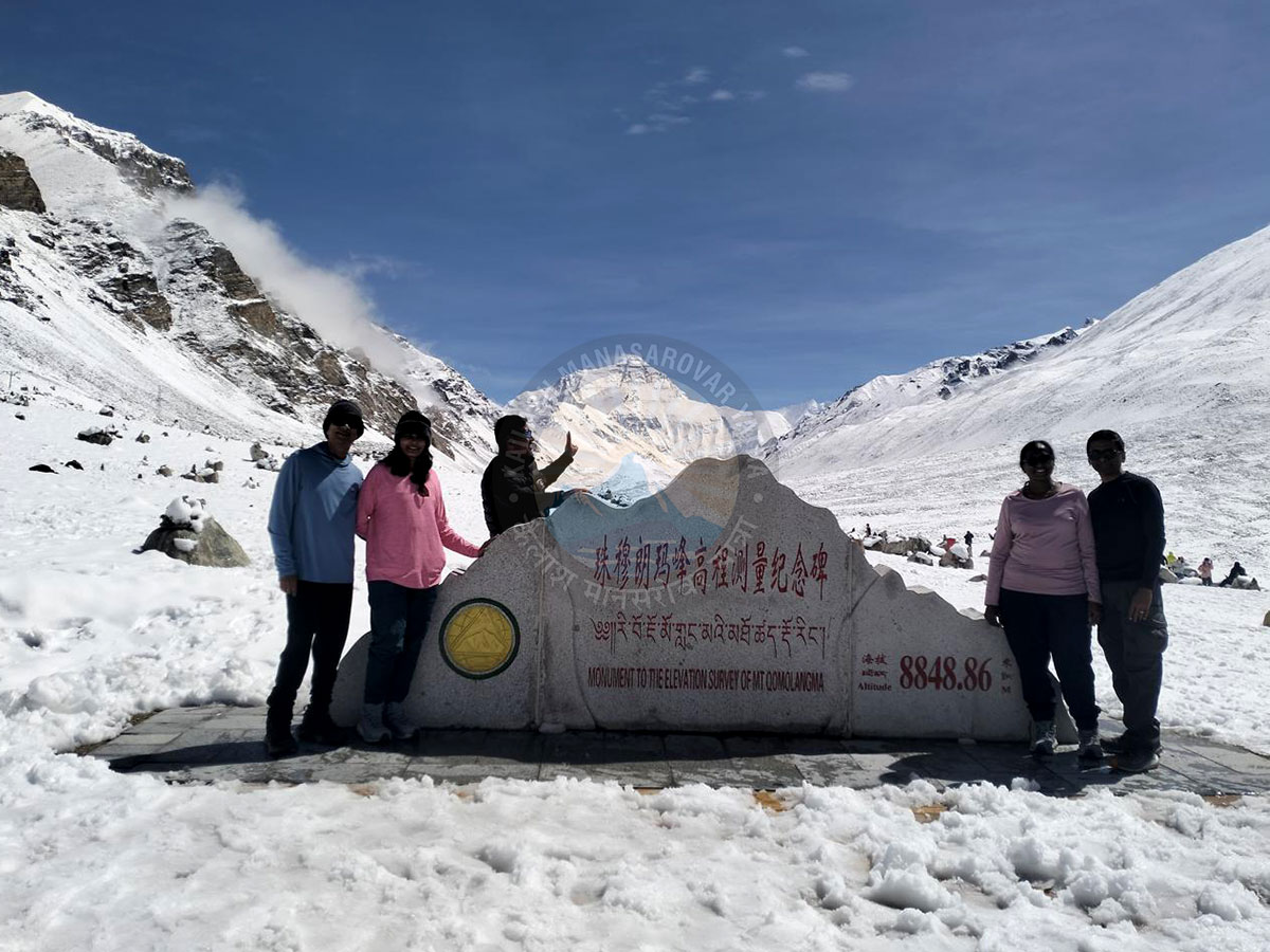 Tourists posing at Mount Everest Base Camp near the 8848.86m altitude monument with snow-covered peaks in the background.