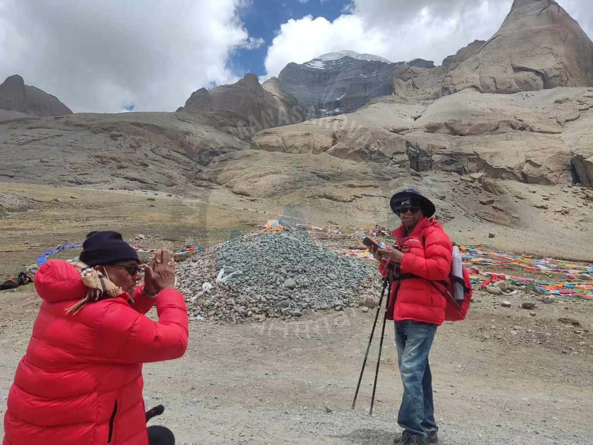 Two pilgrims in red jackets performing parikrama and praying near Mount Kailash with sacred prayer flags and rocky terrain in the background.