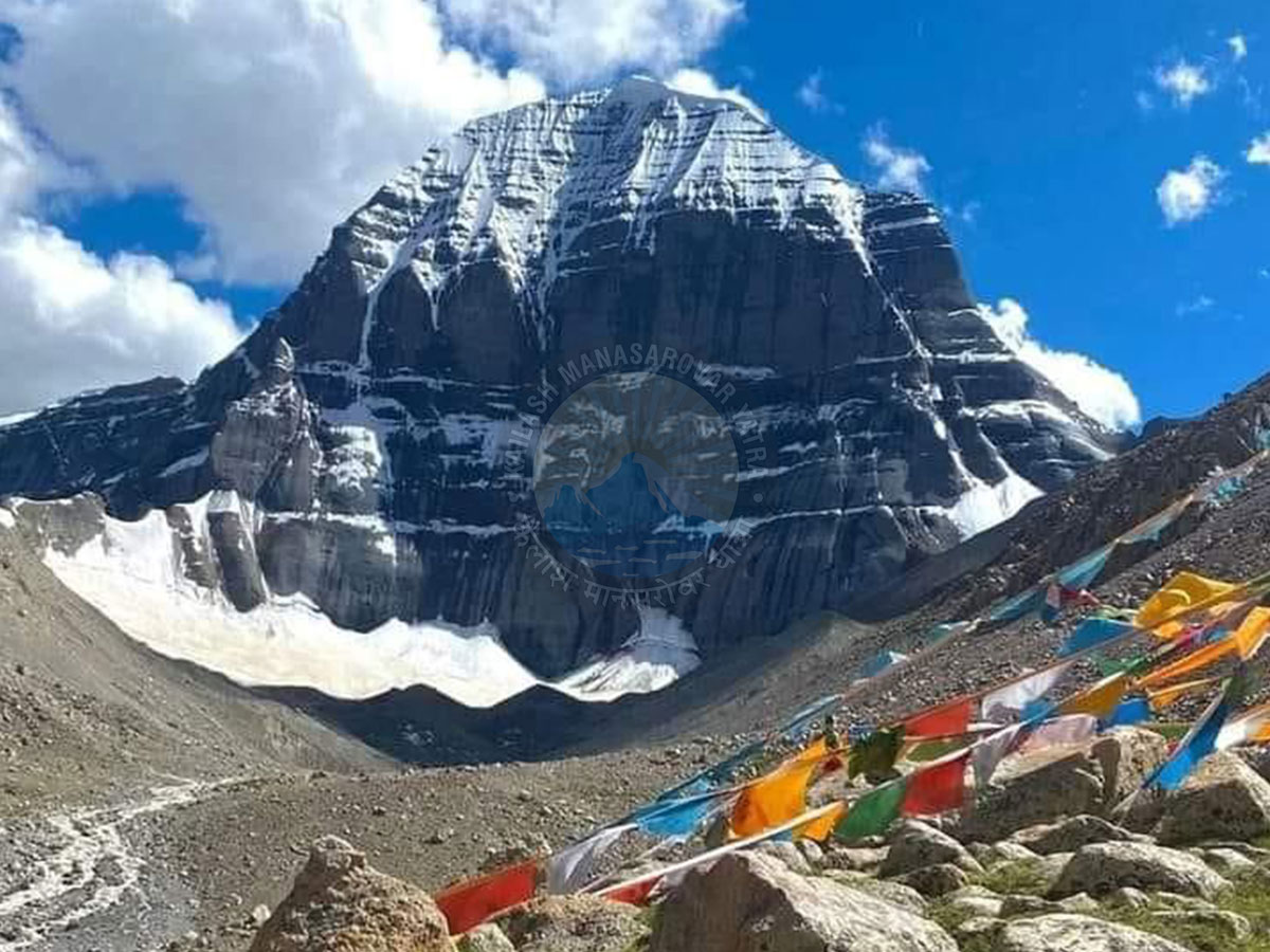 Majestic Mount Kailash peak with snow-covered ridges under a blue sky, adorned with colorful Tibetan prayer flags in the foreground.