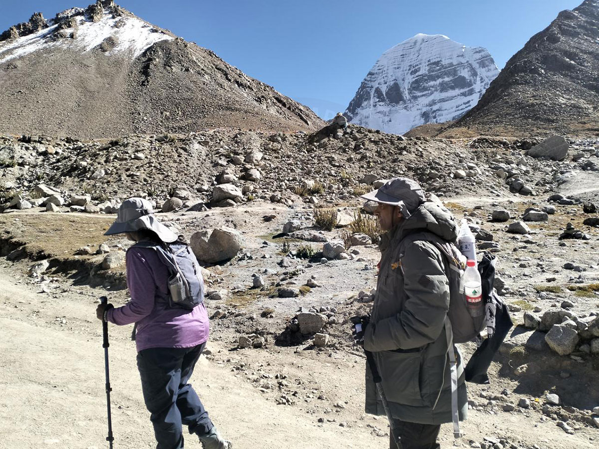 Two hikers trekking on a rugged mountain trail with Mount Kailash in the background, during a high-altitude pilgrimage journey.