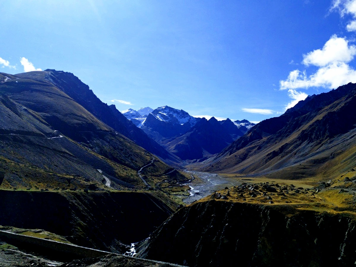 A photograph of the Himalayas with the snowy Om Parvat peak visible in the distance. The peak is surrounded by other mountains, with a river flowing through a valley below. The sky is a clear blue with some clouds.